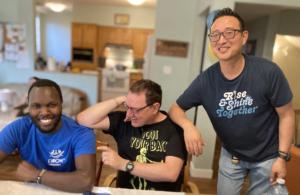 Three men, of diverse races and abilities, with arms leaning on each other and smiling at a dining room table.